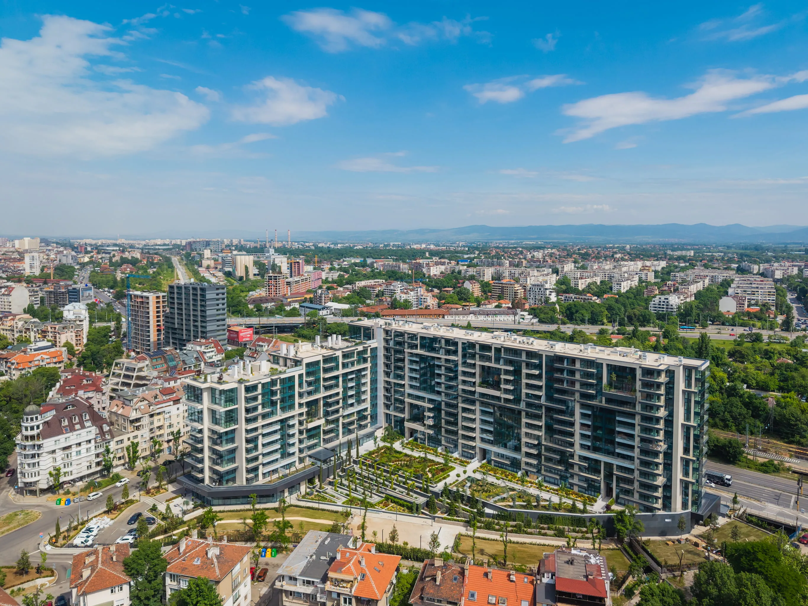 Aerial view of large modern apartment buildings with gardens in a city under a blue sky.